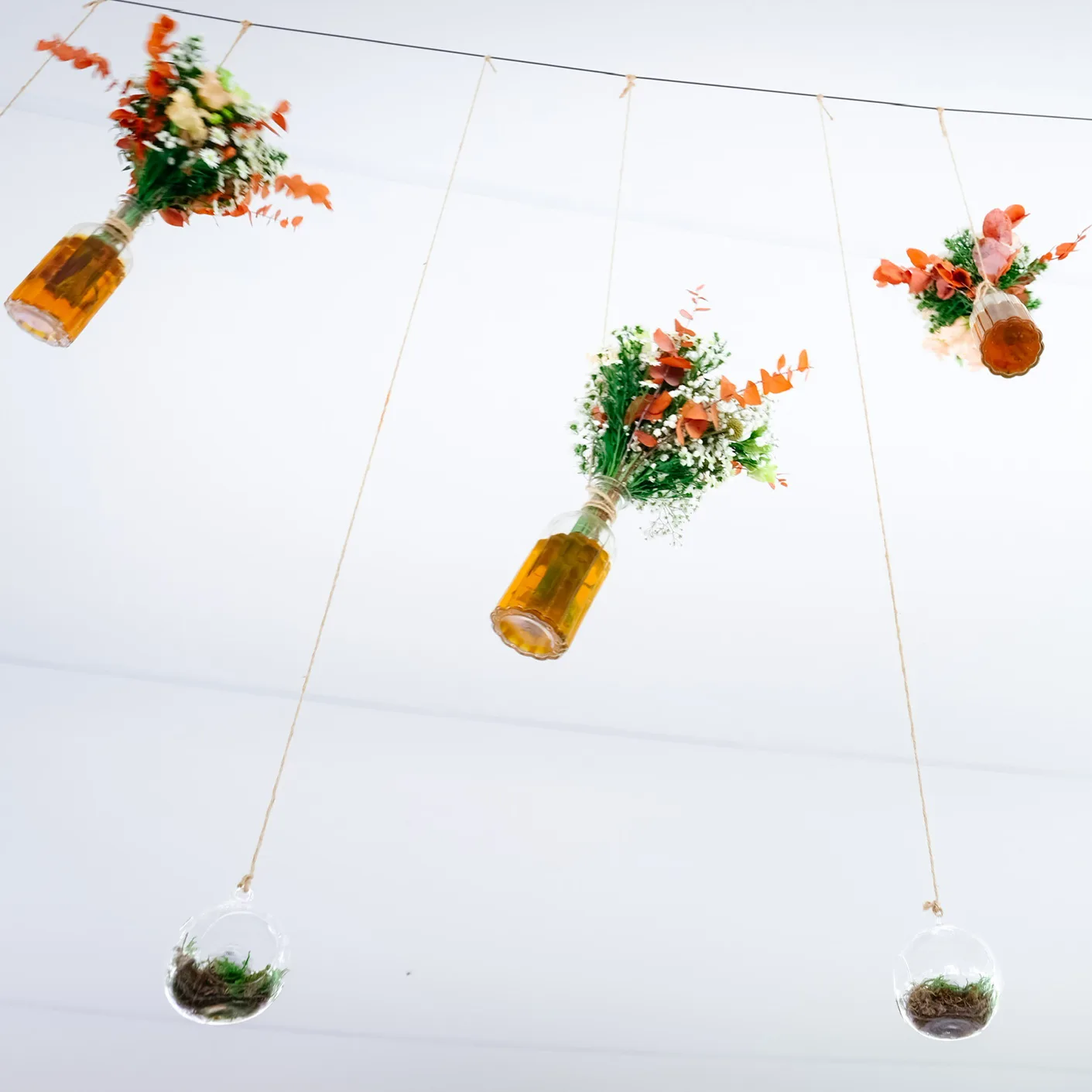 Décoration florale aérienne pour mariage, bouquets suspendus dans des bouteilles ambrées et sphères transparentes garnies de mousse, créant une ambiance légère et naturelle