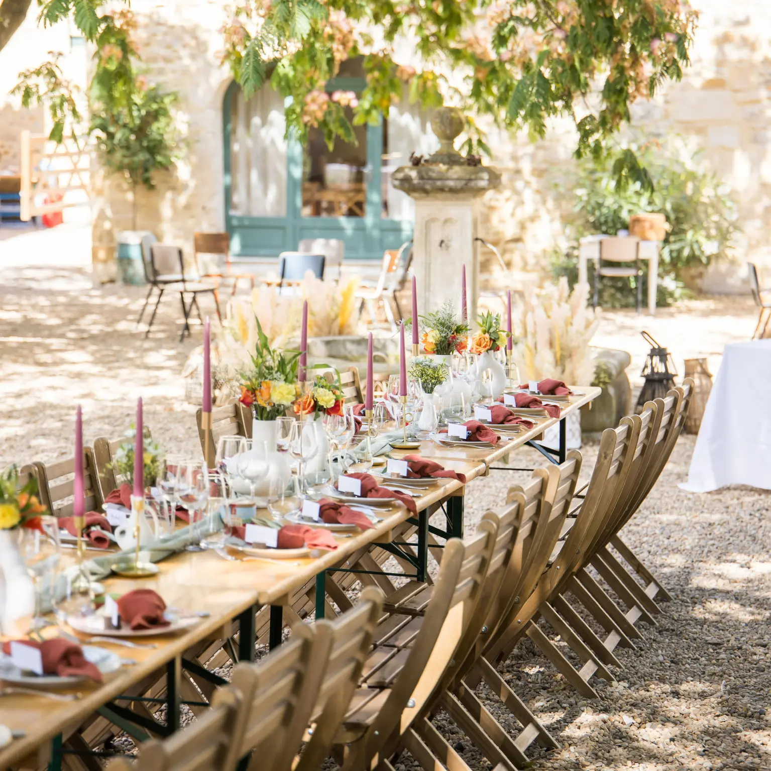 Table de réception de mariage installée en plein air dans une cour en pierre, décorée de fleurs colorées, de bougies mauves et de serviettes bordeaux pour une ambiance élégante et naturelle