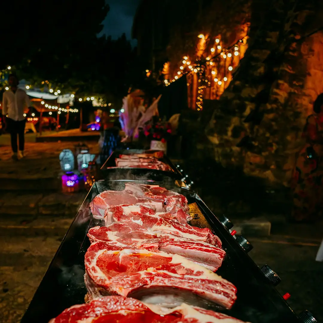 Grandes pièces de viande en train de griller sur plancha en extérieur, ambiance nocturne avec guirlandes, par Yello Event dans le Gard