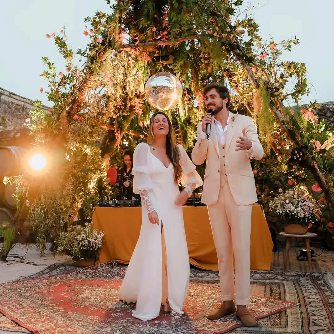 Un couple de mariés sous une arche florale ornée de verdure et d’un miroir disco, durant leur discours de mariage, dans une ambiance chaleureuse et festive.