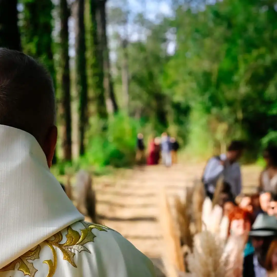 Vue de dos d’un officiant en robe blanche brodée regardant une allée de cérémonie de mariage en pleine forêt.