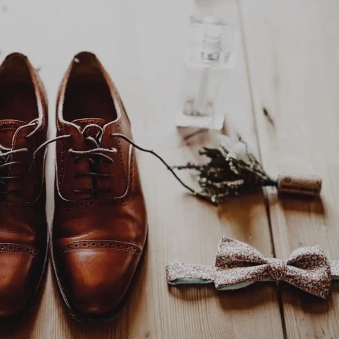 Accessoires de mariage pour homme disposés sur un plancher en bois : chaussures en cuir, nœud papillon fleuri, boutonnière et parfum.