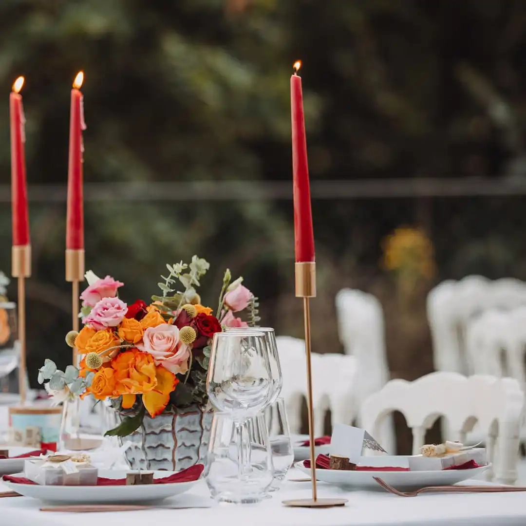 Centre de table floral coloré avec des bougies rouges allumées sur une table de réception extérieure.