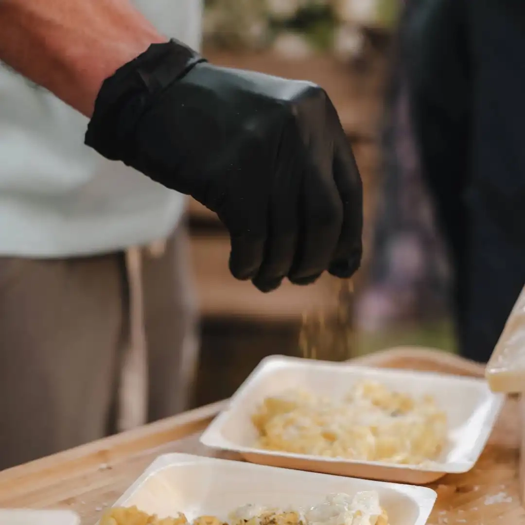 Main gantée en noir saupoudrant un plat de pâtes dans une assiette en carton lors d’un service traiteur.